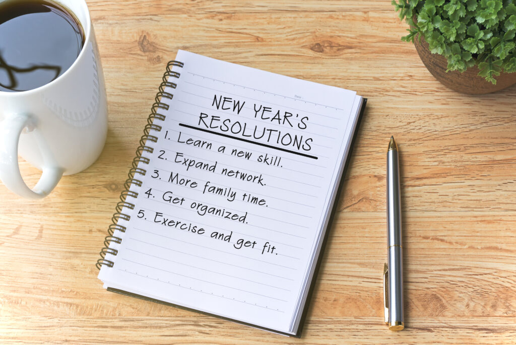 New year's Resolutions written on Notepad with Pen, Coffee and Plant on Top of Wooden Table.